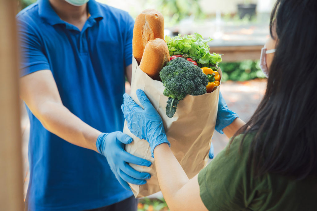 A man wearing protective gloves and a mask delivers groceries to a woman also wearing a face mask and gloves.